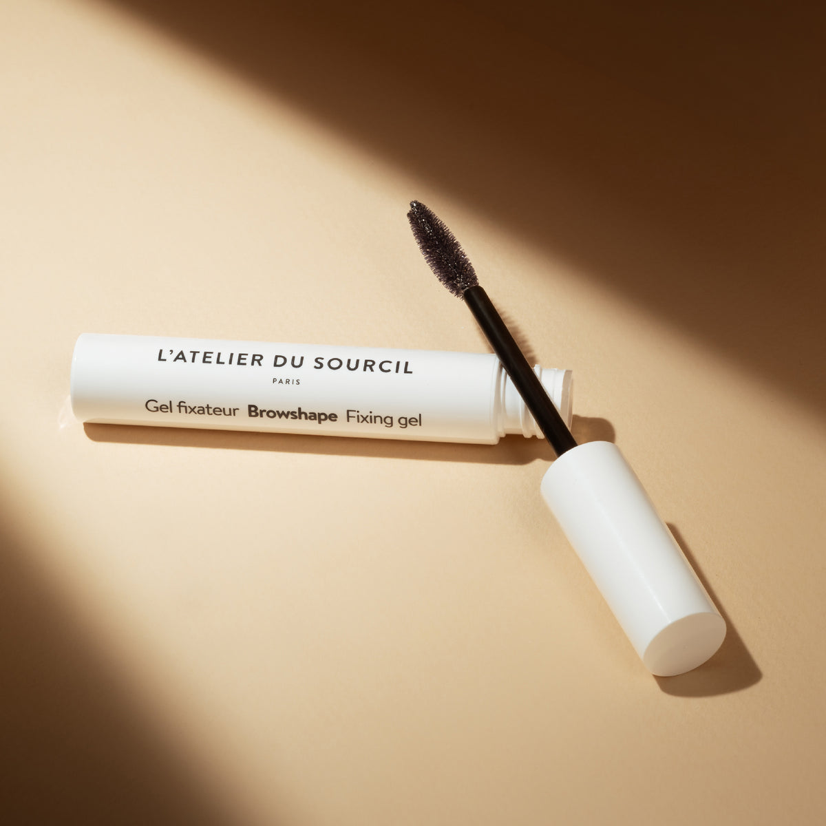 Browshape transparent ouvert, tube blanc et brosse dans un rai de lumière sur fond beige, gel fixateur L'Atelier du Sourcil