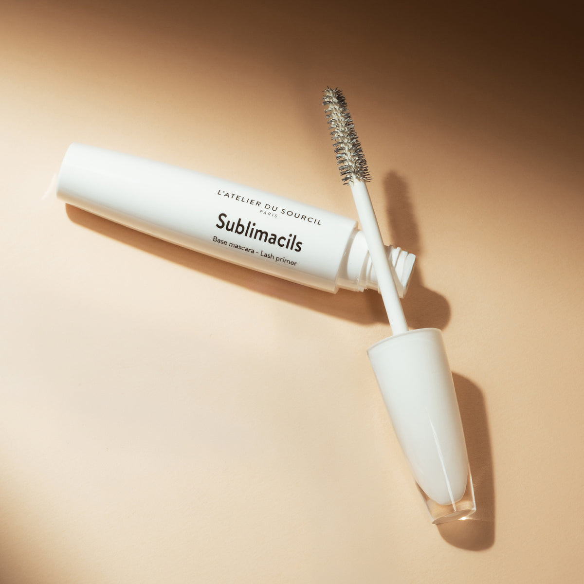 Sublimacils ouvert, brosse posée sur le flacon blanc, sur fond beige en lumière chaude, base mascara L'Atelier du Sourcil