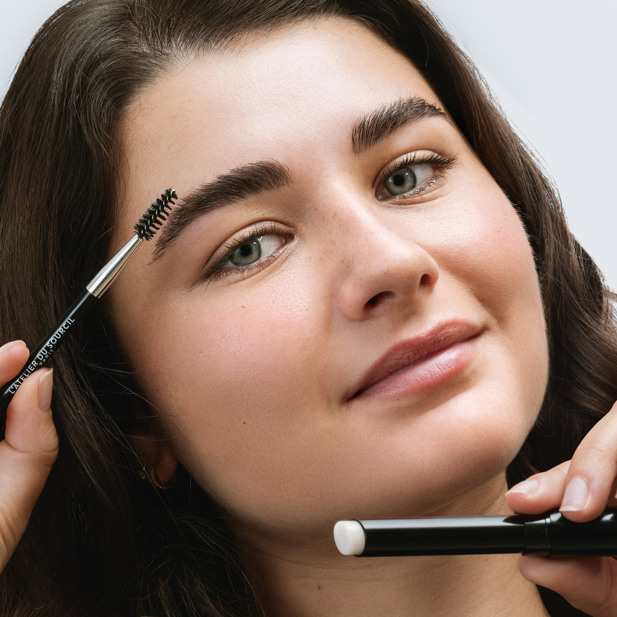 Modèle brune se brossant le sourcil au goupillon, Browstyler tenu près du visage, cire sourcils L'Atelier du Sourcil