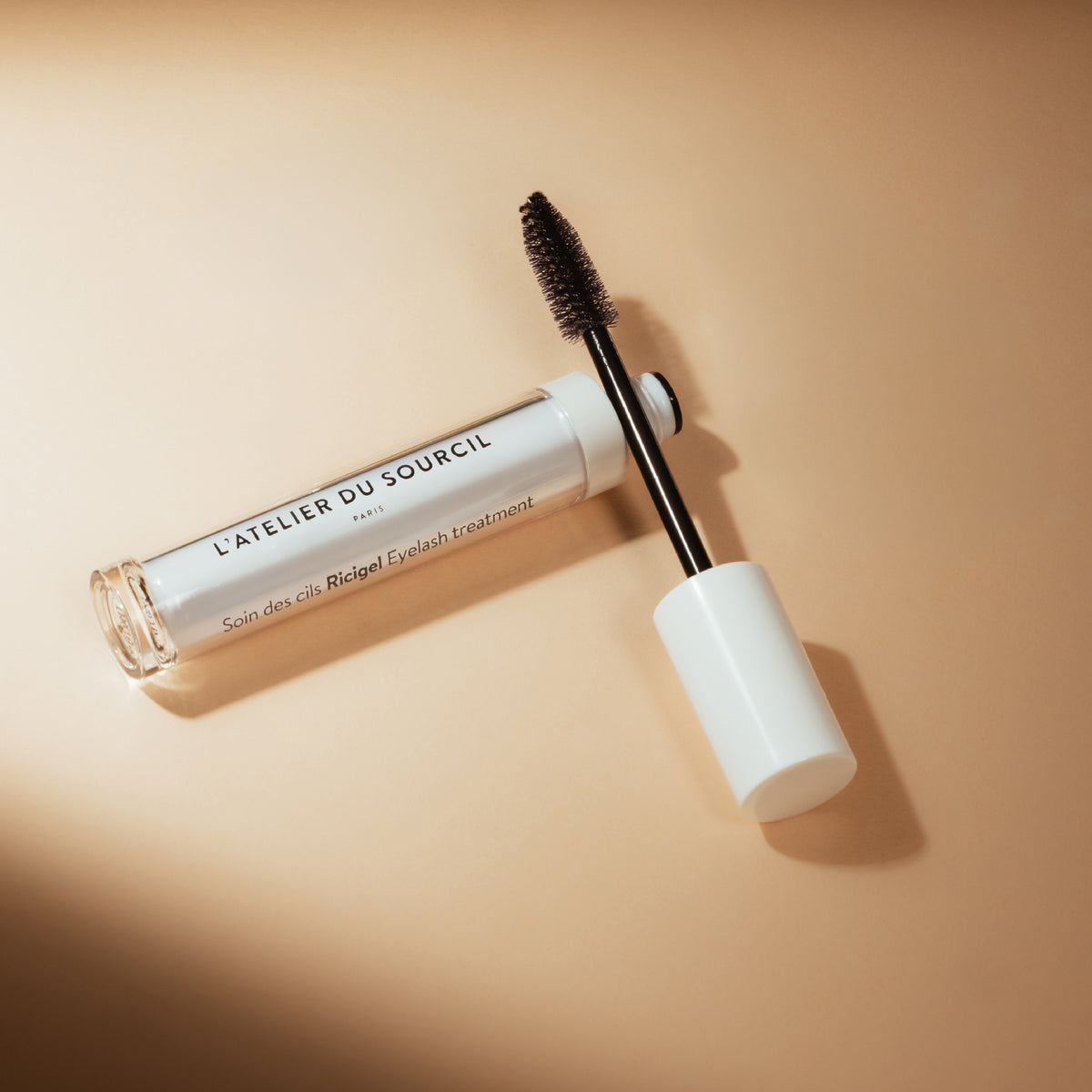 Ricigel ouvert, brosse posée en travers du flacon sur fond beige en lumière chaude, soin des cils L'Atelier du Sourcil