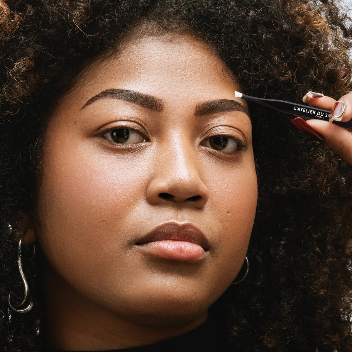 Épilation du bout du sourcil avec le Stylbrow par une modèle aux cheveux bouclés, pince à épiler L'Atelier du Sourcil